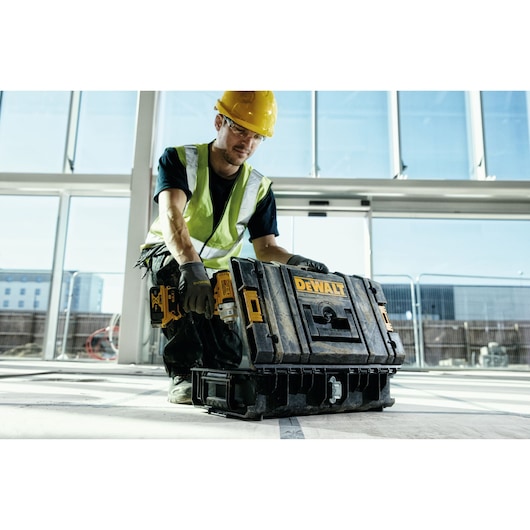 A worker in a safety vest and hard hat uses a DEWALT tool box on a construction site indoors. The worker is kneeling and handling the toolbox, which is placed on the floor.