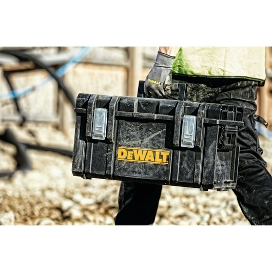 A person wearing work gloves is carrying a DEWALT-branded black toolbox on a construction site.