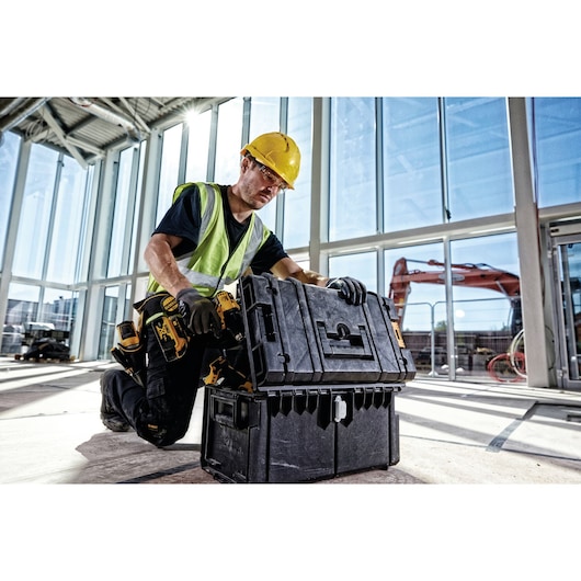 A construction worker wearing a yellow hard hat, gloves, and a reflective vest is kneeling indoors on a sunlit construction site. They are handling a large black DEWALT toolbox (SKU: 1-70-322) with organized compartments, stacked on another toolbox. There is construction equipment and large windows in the background.