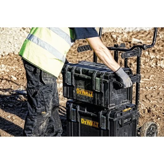 A person wearing a reflective safety vest and gloves is handling stacked DEWALT toolboxes on a wheeled cart at a construction site.