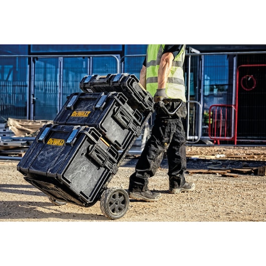 A person in work clothes and a safety vest is pulling a stack of three black DEWALT tool storage boxes on a wheeled trolley at a construction site.