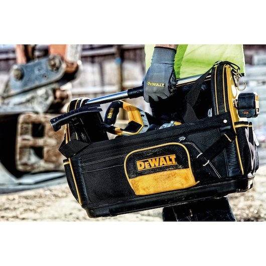 A DEWALT branded tool bag being carried by a gloved hand, containing various tools including a hammer and power tool, with construction equipment visible in the background.