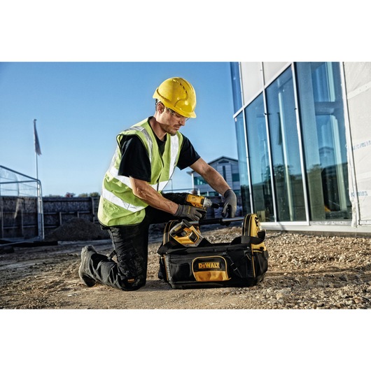 A person wearing a yellow hard hat and safety vest is kneeling outdoors at a construction site next to a DEWALT branded tool bag, holding a power tool.