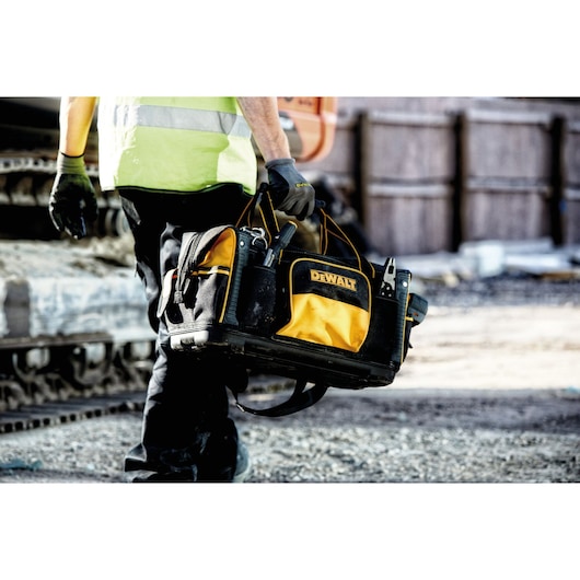 A person wearing work gloves and a high-visibility vest is carrying a DEWALT tool bag at a construction site. The black and yellow tool bag is filled with various tools.