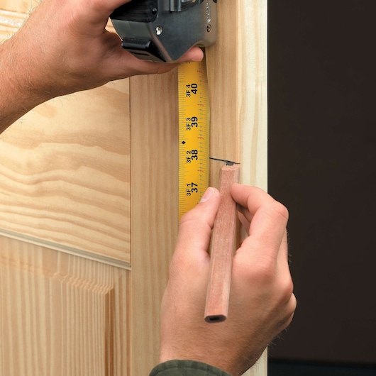A close-up of hands using a yellow DEWALT tape measure to measure the height on a wooden door frame, with a pencil marking the measurement.