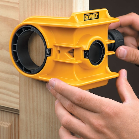 A yellow DEWALT door lock installation kit is being used to mark or cut a hole in a wooden door. The tool is held firmly against the door by a person's hands, showing the process of preparing for lock installation.
