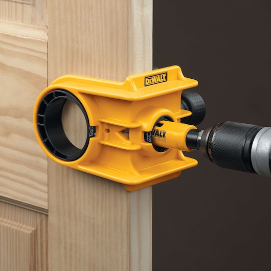 A yellow DEWALT door lock installation kit is being used to drill a hole in a wooden door. The kit is attached to the edge of the door and a drill is inserted into the guide.