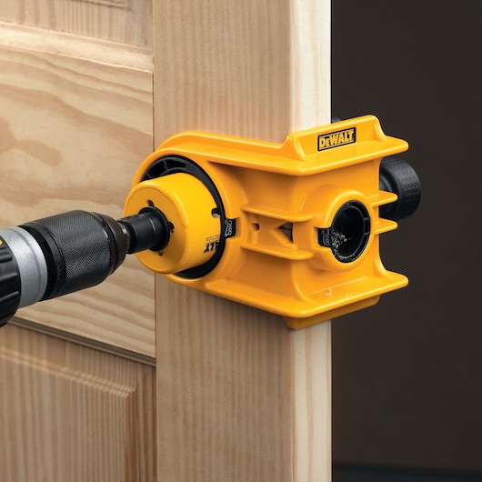 A DEWALT yellow hole saw kit is attached to a wooden door edge, being used with a power drill to create a hole. The tool is designed for precise door hardware installation.