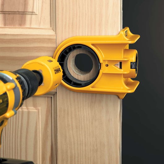 A DEWALT yellow door lock installation kit is being used with a drill to create a circular hole in a wooden door.