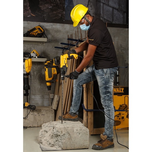 A person wearing a yellow safety helmet and gloves operates a DEWALT electric rotary hammer drill on a concrete block in an indoor workspace. There are other DEWALT tools and a tool case visible in the background.