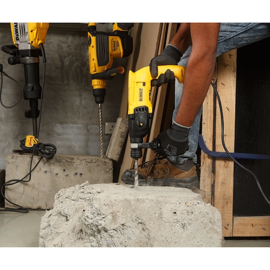 A person wearing gloves and work boots uses a yellow DEWALT hammer drill to drill into a concrete slab. Other DEWALT power tools are visible in the background in a workshop setting.