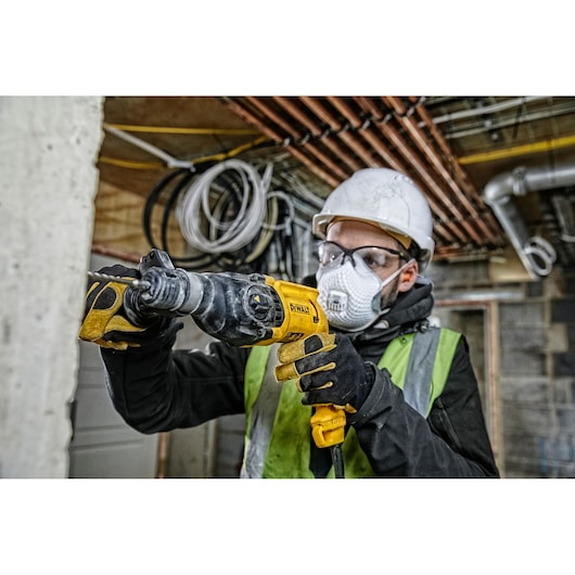 A person wearing a safety helmet and vest is using a DEWALT D25033K power tool on a construction site. The individual’s face is blurred for privacy. The background features exposed pipes and wires.