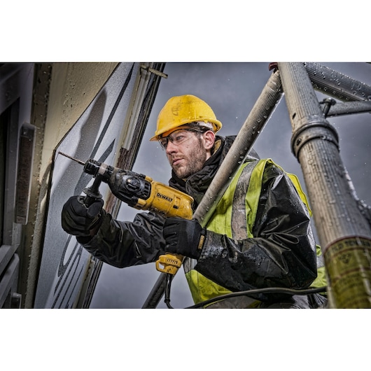 A construction worker wearing a yellow hard hat and reflective vest uses a DEWALT D25033K power drill to work on a wall, standing on scaffolding outdoors in rainy conditions. The person's face is blurred for privacy.