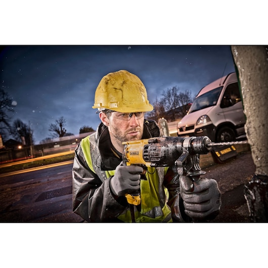 A person wearing a yellow hard hat and reflective safety vest uses a DEWALT electric drill outdoors, with a van and blurred lights visible in the background. The person's face is obscured.