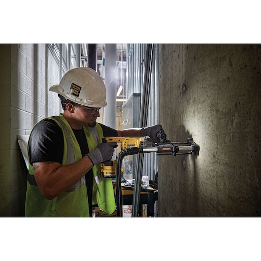 A construction worker wearing a hard hat and safety vest uses a DEWALT power tool with a dust extraction attachment to drill into a concrete wall at an indoor jobsite. The worker's face is blurred.
