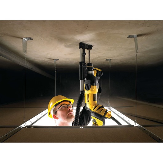 A person wearing a yellow safety helmet and gloves is using a DEWALT D25301D power tool to drill into a ceiling, seen from below through an open ceiling panel. The face of the person is blurred for privacy.
