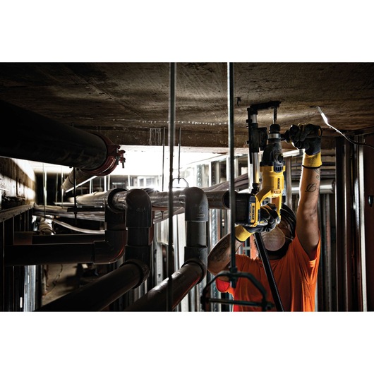 A worker wearing safety gloves and an orange shirt uses a yellow DEWALT power tool to drill into a concrete ceiling in an industrial setting with pipes and metal frameworks.