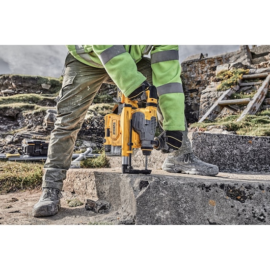 A person wearing work clothes and safety gloves is using a yellow DEWALT rotary hammer drill on a concrete surface outdoors. The drill is equipped with a dust collection attachment.