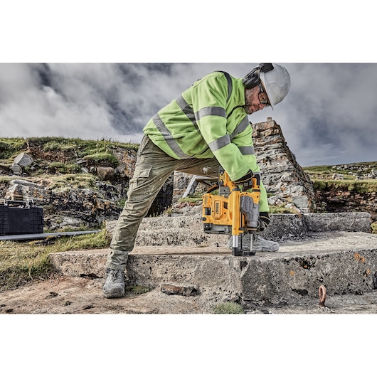 A construction worker wearing a green reflective jacket, protective helmet, and ear protection is using a yellow DEWALT drill on a concrete surface outdoors.