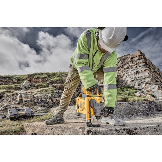 A worker wearing a hard hat and high-visibility jacket operates a yellow DEWALT rotary hammer drill on a concrete surface outdoors, with rocky terrain and a tool case visible in the background.