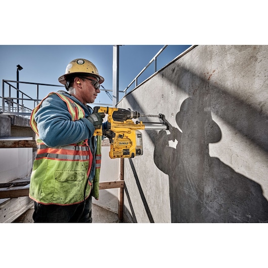 A construction worker wearing a helmet and safety vest is using a DEWALT D25333K rotary hammer drill to bore into a concrete wall at an outdoor construction site.