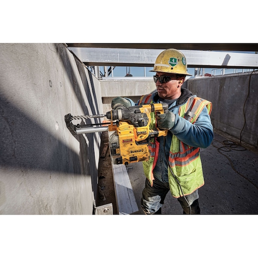 A construction worker wearing a yellow hard hat and reflective safety vest uses a DEWALT rotary hammer drill to drill into a concrete wall at a construction site.