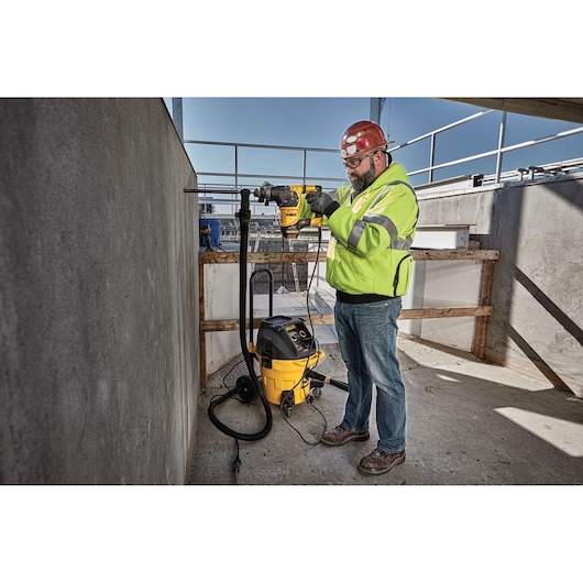 A person wearing a high-visibility jacket and a red hard hat is using a DEWALT power tool to drill into a concrete wall at a construction site. A yellow and black DEWALT vacuum is connected to the tool to manage dust.