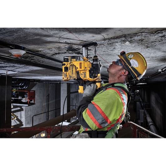 A construction worker, wearing a safety helmet and high-visibility clothing, is using a yellow DEWALT rotary hammer drill to drill into a concrete ceiling at a work site.