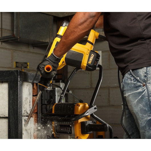 A person using a DEWALT rotary hammer drill to chisel concrete in an industrial setting. Dust and debris are visible around the tool during operation.