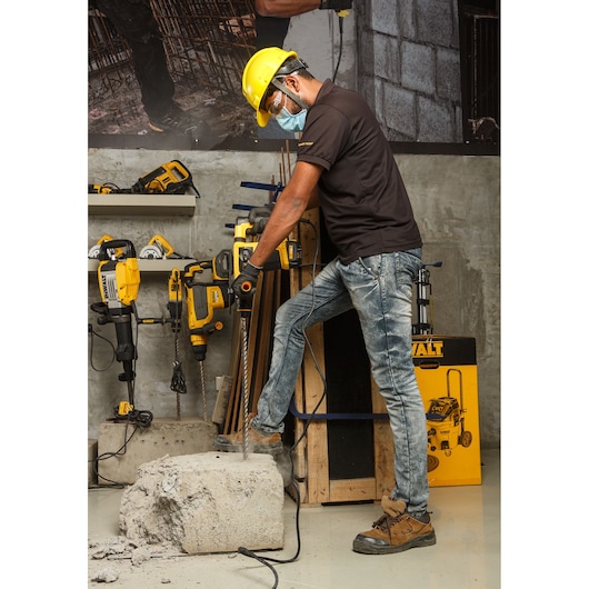 A person wearing safety gear uses a DEWALT hammer drill to break a large concrete block in a workshop. Other DEWALT power tools are visible in the background, along with construction materials.