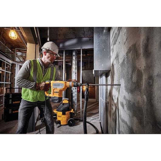 A worker wearing a safety helmet and vest operates a DEWALT rotary hammer drill (SKU: D25614K) to drill into a concrete wall at a construction site. DEWALT equipment and tools are visible in the background.