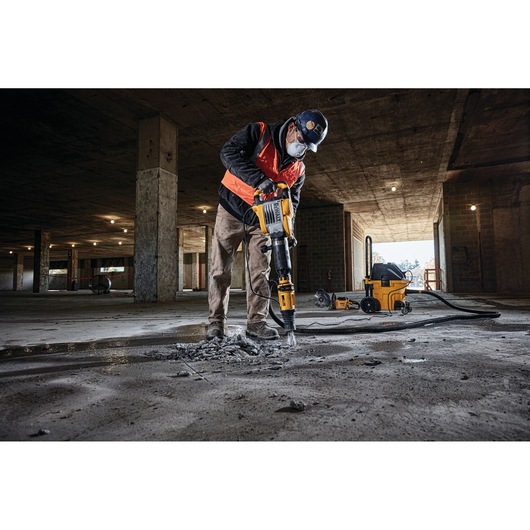 A worker in protective gear using a yellow DEWALT jackhammer to break concrete on the floor of a large, partially constructed building. A DEWALT vacuum machine is visible in the background.