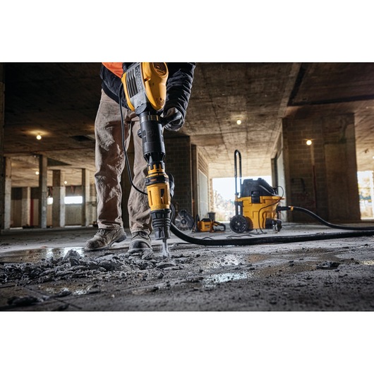 A worker using a yellow DEWALT demolition hammer to break up concrete on the floor of a construction site. There is dust and rubble on the ground, and other yellow DEWALT tools are visible in the background.