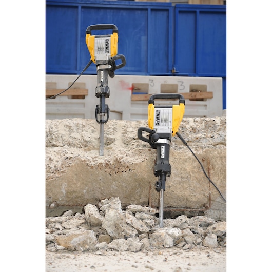 Two DEWALT electric demolition jackhammers are shown breaking up a large concrete slab at a construction site, with rubble scattered on the ground.