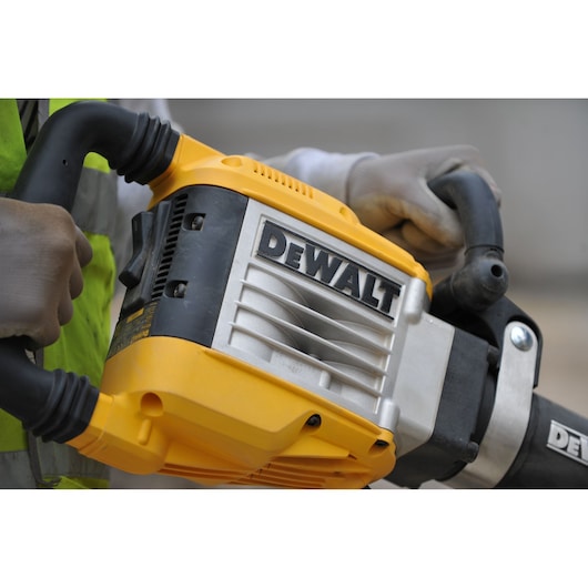 Close-up of a person holding a DEWALT D25961K demolition hammer, wearing gloves and a safety vest.