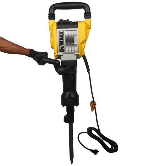 A person wearing a glove is holding a DEWALT electric demolition hammer with a pointed chisel bit and a long power cord attached.