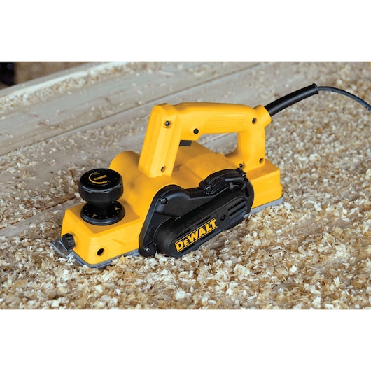 A DEWALT electric hand planer placed on a wooden surface covered with wood shavings. The planer is yellow and black, and is plugged in with a visible power cord.