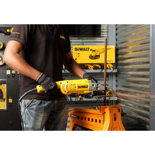 A person wearing gloves and a black shirt uses a yellow DEWALT angle grinder to cut a metal rod, producing sparks. The workspace includes a sturdy orange metal table and DEWALT products and tools displayed in the background.