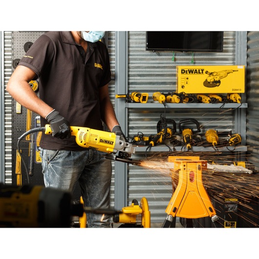 A person wearing a black shirt, gloves, and a face mask is using a DEWALT D28413 angle grinder to cut metal, creating sparks. The scene is set in a workshop with various DEWALT tools and equipment visible in the background.
