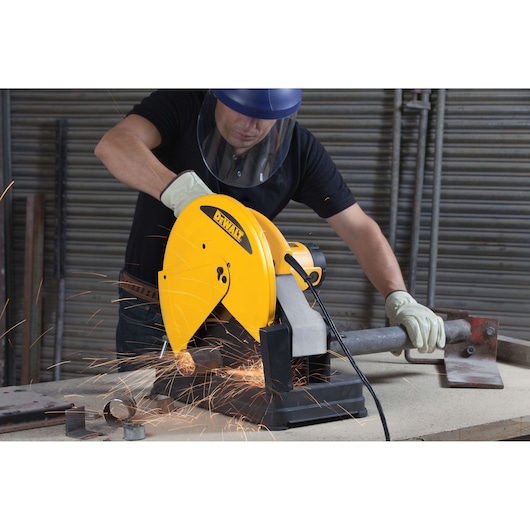 A person wearing protective gloves and a face shield is using a DEWALT D28720 yellow metal cutting saw, generating sparks, to cut a metal pipe on a workbench in an industrial setting.