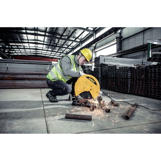 A worker wearing a safety helmet and reflective vest is operating a DEWALT D28730 metal cutting saw, producing sparks as it cuts through metal pipes in an industrial warehouse.