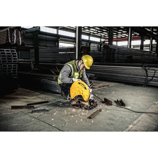 A person wearing a yellow safety helmet and reflective vest uses a DEWALT metal cutting saw to cut steel bars in an industrial warehouse. Sparks are visible during the cutting process.