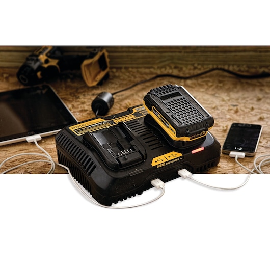 A DEWALT DCB102 dual battery charger on a workbench, charging a DEWALT battery and simultaneously powering two electronic devices, a tablet and a smartphone, using USB ports. A cordless power tool is in the background.
