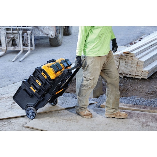 A worker wearing gloves and work clothes pulls a rolling cart loaded with DEWALT toolboxes and battery chargers on a construction site. Wooden planks and building materials are visible in the background.
