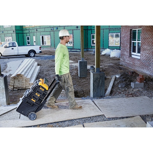 A construction worker wearing a hard hat and green shirt pulls a rolling cart stacked with black and yellow DEWALT toolboxes across a construction site.