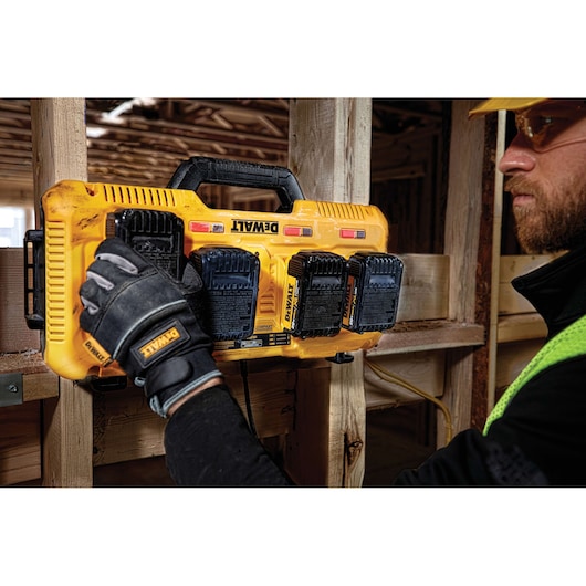 A Dewalt DCB104 yellow multiport battery charger mounted on a wooden beam at a construction site. A person wearing Dewalt-branded gloves is handling the charger, and there are four Dewalt batteries attached to it.