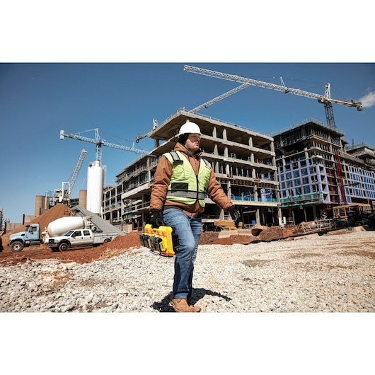 A construction worker wearing a safety vest and hard hat is carrying a yellow DEWALT DCB104 battery charger with batteries at a construction site. Several cranes and unfinished buildings are visible in the background.