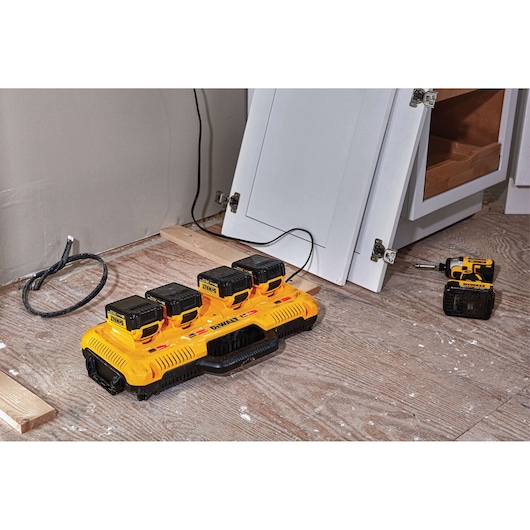 A DEWALT DCB104 battery charger with four batteries is placed on a wooden floor in a workshop. Nearby, a DEWALT cordless tool and cabinet doors are visible, with some construction materials scattered around.