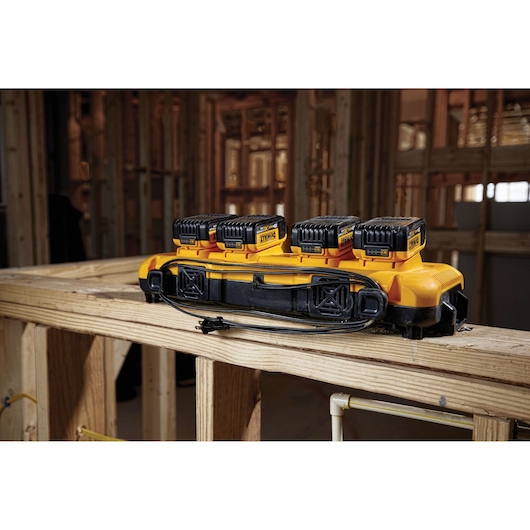 A DEWALT DCB104 yellow four-port battery charger with four DEWALT batteries, placed on a wooden beam in an unfinished construction site.