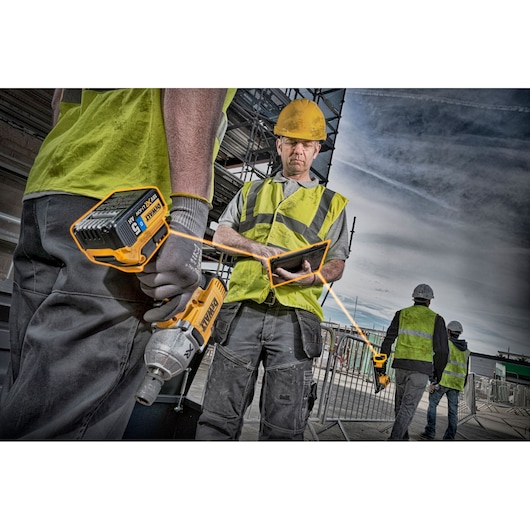 Construction workers wearing safety vests and helmets are on an outdoor building site. One worker holds a DEWALT power tool with a DCB184 battery, while another checks a tablet. In the background, two more workers walk near scaffolding.